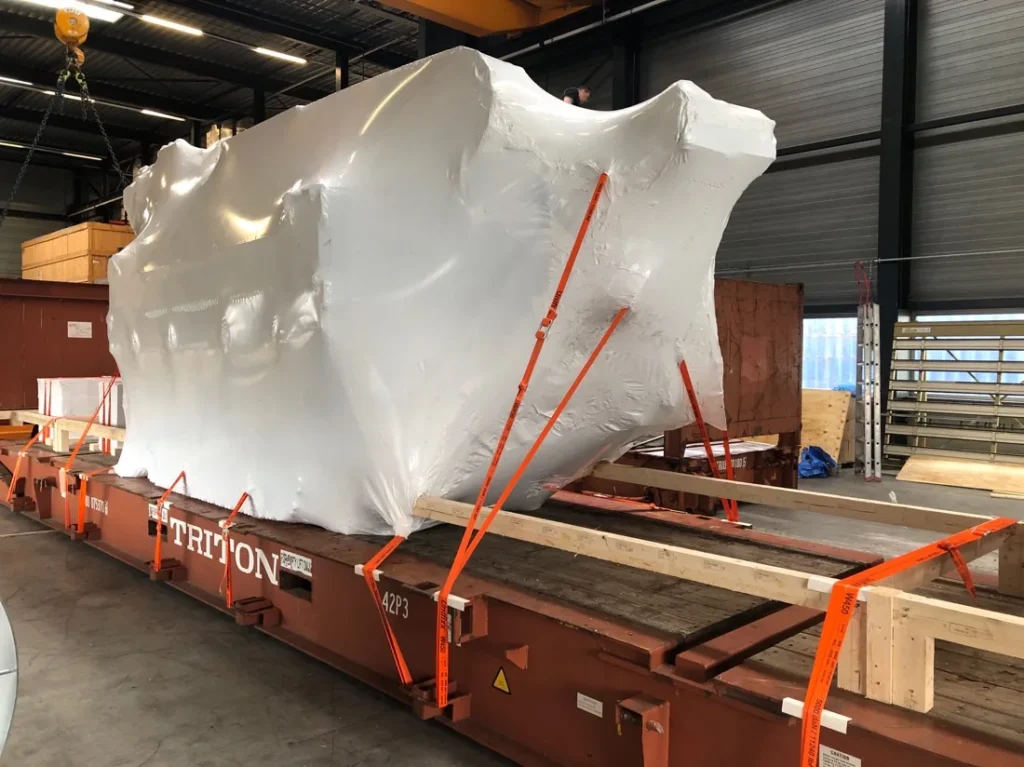 In folie verpakte machine met oranje sjorbanden op een flatrack in de loods
