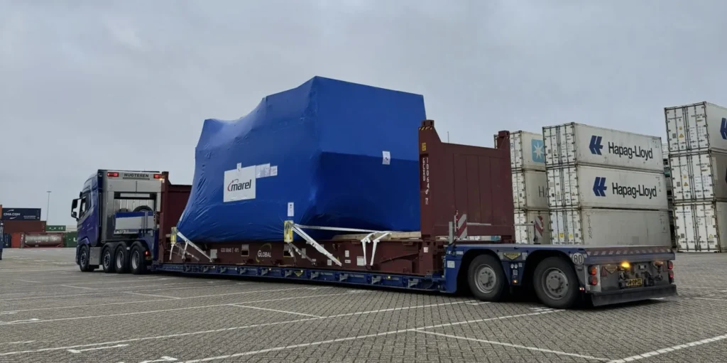 Dieplader met een in folie verpakte machine tussen zeecontainers